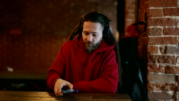 Hipster is Listening to Music By Headphones and Smartphone in Cafe Stylish Man with Dreadlocks