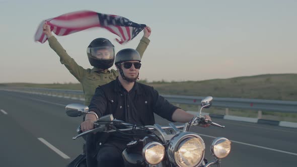 Man in Helmet and Black Glasses Rides with Woman Holding an American Flag Flying High in the Wind on