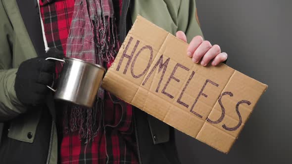 Man in dirty clothes holds a cardboard sign with the inscription homeless and begging with a cup.