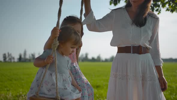 Mom Shakes Two Small Sisters 1 and 3 Years Old on a Wooden Swing with Rope Handles