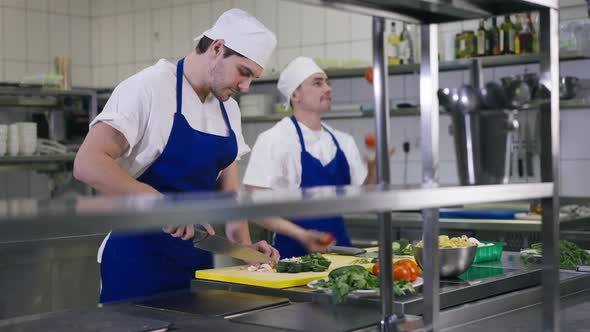 Focused Man Cutting Salad Ingredients As Cheerful Colleague Juggling Tomatos at Background