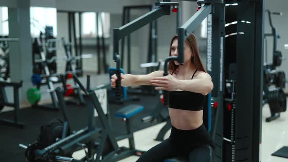 A pretty woman with flowing hair is sitting and doing exercise for the chest in the gym.