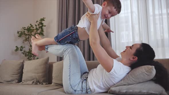 Happy Healthy Family Young Mom Lifting Cute Little Child Son Up Playing Plane on Sofa at Home, Funny