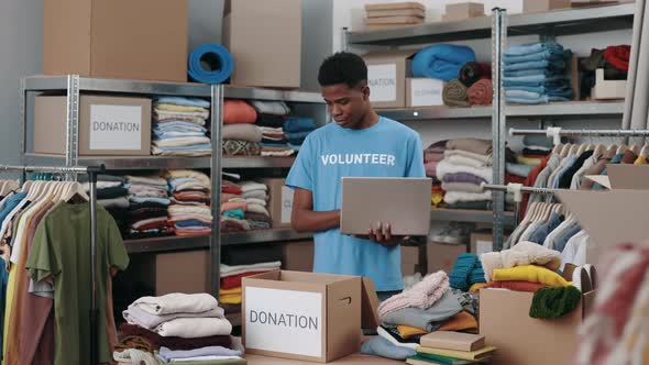 Multiracial Male Volunteer Using Laptop While Working at the Donating Center with a Lot of Cupboard