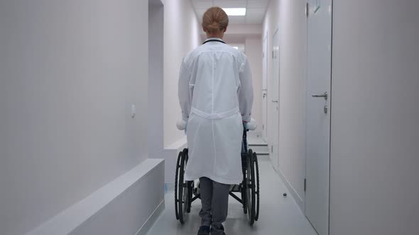 Back View Doctor Pushing Wheelchair with Boy Along Hospital Corridor in Slow Motion