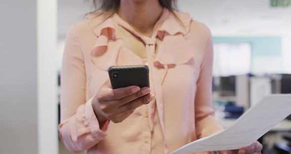 Video of midsection of biracial businesswoman using smartphone and walking in office