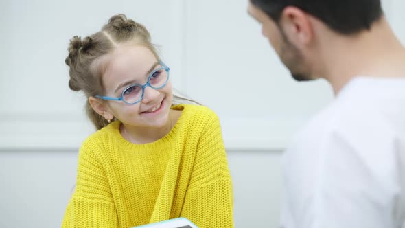 Interested Pretty Caucasian Girl in Eyeglasses Listening to Blurred Doctor with Tablet Talking