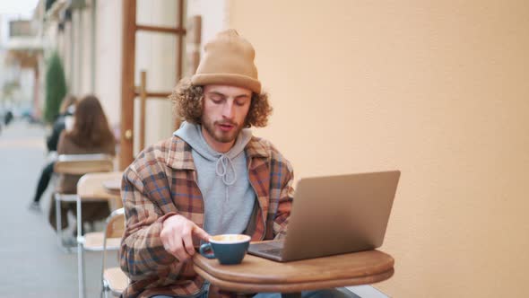 Handsome curly-haired man texting by phone and drinking coffee in cafe