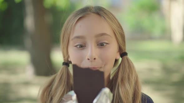 Close-up Portrait of Joyful Little Girl Eating Tasty Chocolate Outdoors. Excited Caucasian Kid