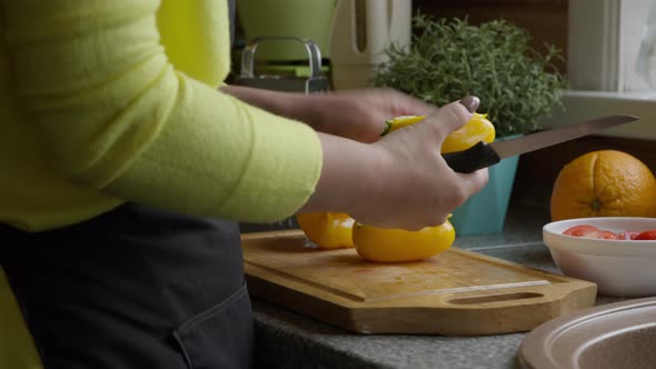 Housewife in Apron Cuts Yellow Sweet Pepper Paprika with Knife on Wooden Board