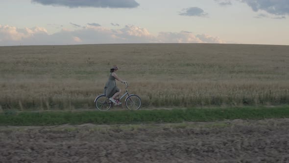 Woman in dress and hat rides pleasure bike along path in wheat field on summer evening