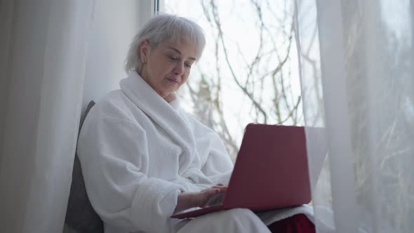 Concentrated Mature Woman Sitting on Windowsill Typing on Laptop Keyboard Surfing Internet