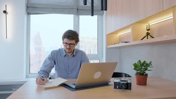 Man in glasses works at a laptop against the background of a window. Freelancer working remotely.