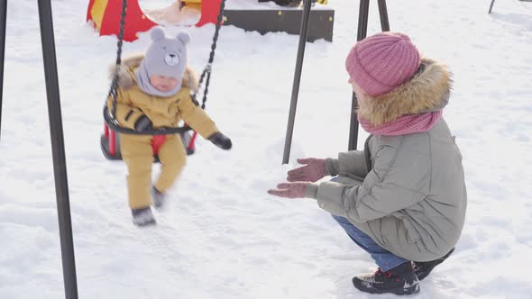 Mom rocks a newborn baby 12-17 months old on a swing on a winter playground