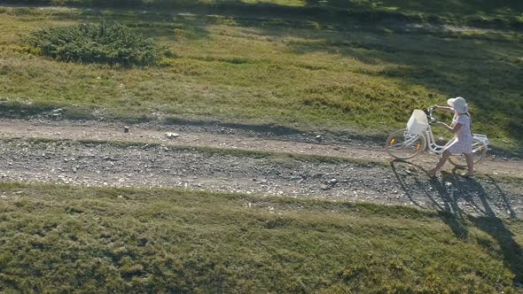 Shot of a Young Womanin a Light Dress and Hat Riding Bicycle on Rural Road