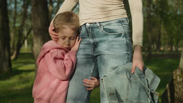 Sleepy Little Girl Leans on Mother Thigh Standing in Park