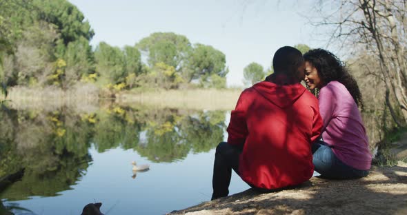 Smiling diverse couple embracing and sitting by lake in countryside