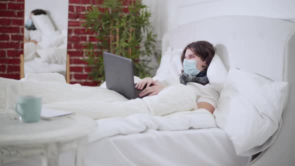 Wide Shot of Ill Tired Young Man in Covid Face Mask Lying in Bed Coughing and Using Video Chat on
