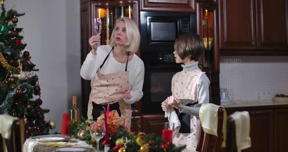 Positive Caucasian Son and Mother in Aprons Setting Christmas Table in Kitchen at Home