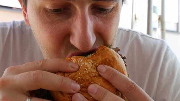 A Young Handsome Man with a Huge Appetite Eats a Big Burger or Cheeseburger on the Terrace of a