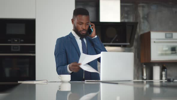 Young African Businessman at Kitchen Counter Working at Home Looking at Report Talking on Cellphone