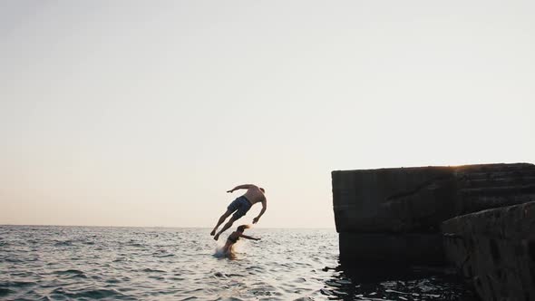 Young Woman and Man Synchronously Doing Trick Jump From a Pier Into the Sea During Beautiful Sunrise