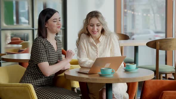 Cheerful Carefree Young Women Surfing Social Media on Laptop and Gossiping Sitting in Cafe Indoors