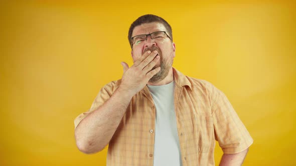 Tired adult male in eyeglasses yawning and demonstrating sleep gesture, bright background