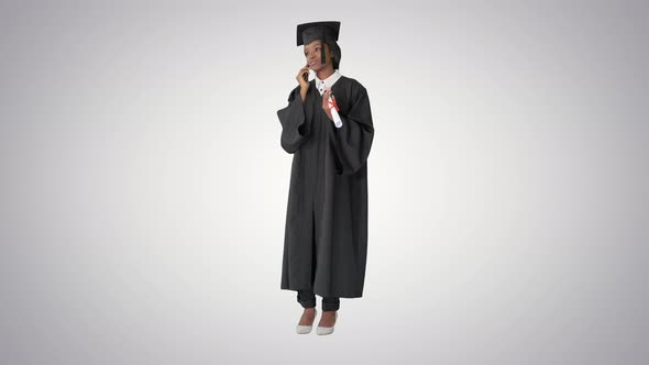 African American Female Student in Graduation Robe Talking on the Phone on Gradient Background