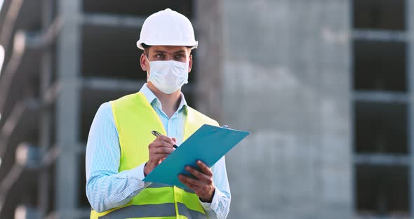 Professional Foreman in Protective Mask and Helmet Checking Building with Plan at Construction Site