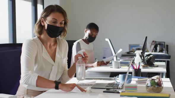 Two diverse female colleagues wearing face mask, sanitizing desk in office