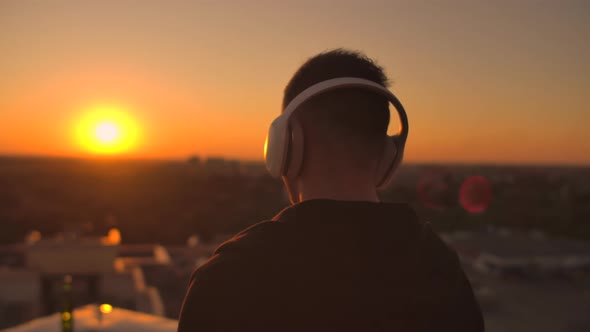 The Camera Is Tracking a Man Walks on the Roof at Sunset with Headphones Looking at the City From