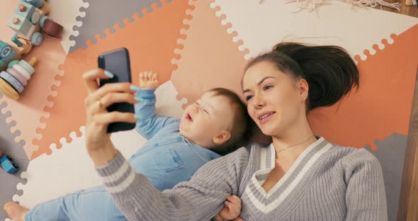 Young Attractive Mom Lies on Floor with Son Dressed in a Blue Bodysuit Boy Looks