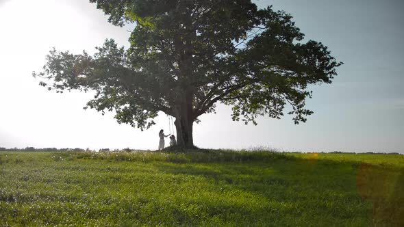 Little Silhouettes of Family Swinging on a Swing on a Branch of Vintage Green Oak Tree.