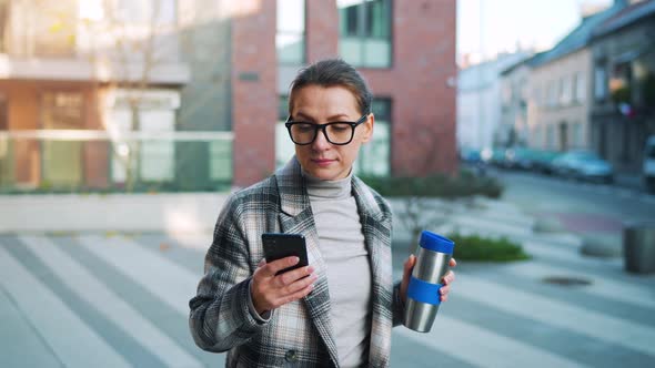 Caucasian Businesswoman with Glasses and a Coat Walks Through the Business District with Thermo Cup