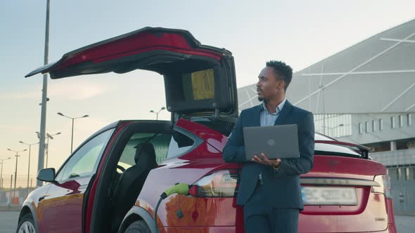 Close Up of African Man in Business Suit Standing Near Charging Electric Car and