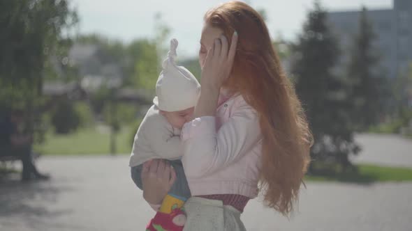 Young Beautiful Redhead Mother Holding a Cute Baby in Her Arms and Talking To Him in a Spring Sunny