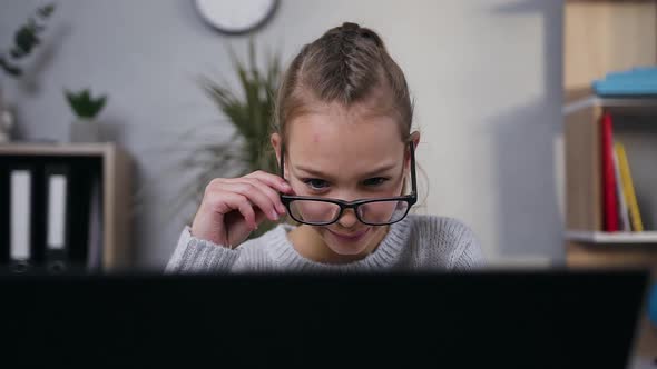 Schoolgirl in Glasses which Sitting in Front of Laptop and Reading the Information on Screen