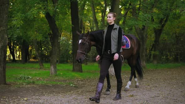 Young Happy Female Horse Rider is Walking with Brown Horse with White Spot on Forehead in Park