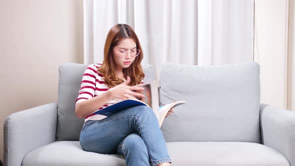 Beautiful young Asian model flipping through picture magazine sitting on the sofa at home.