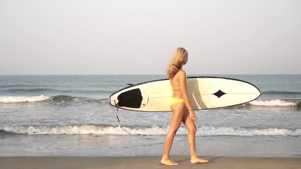 Young Slender Woman Surfer in a Bikini Goes with a Surfboard on the Background of the Sea