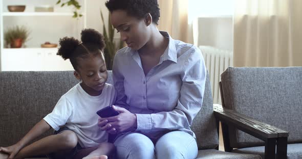 Curly African American Mother and Little Girl Sitting at Home Sofa. Woman Showing Something To Small