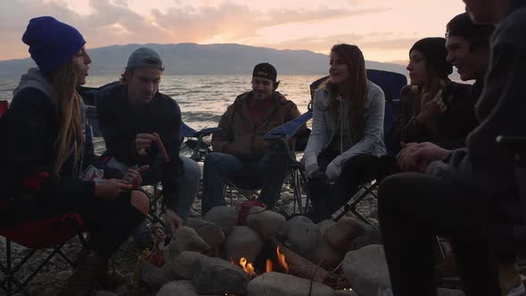 Group of millenials gathered around campfire at sunset