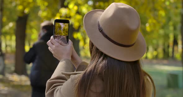 Back View of Young Caucasian Girl in Brown Hat Taking Photos of Autumn Park on Her Smart Phone