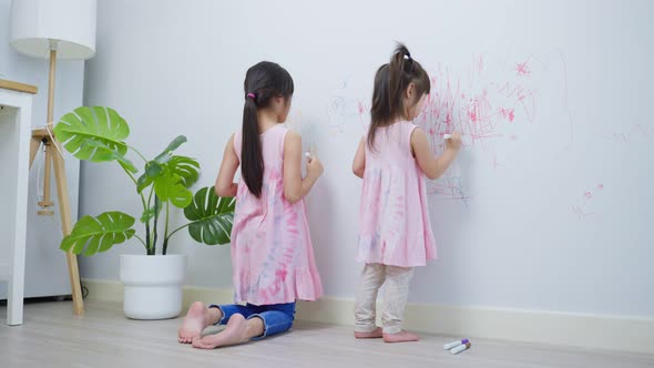 Asian young sibling kid girl having fun drawing and coloring on white wall in living room at home.