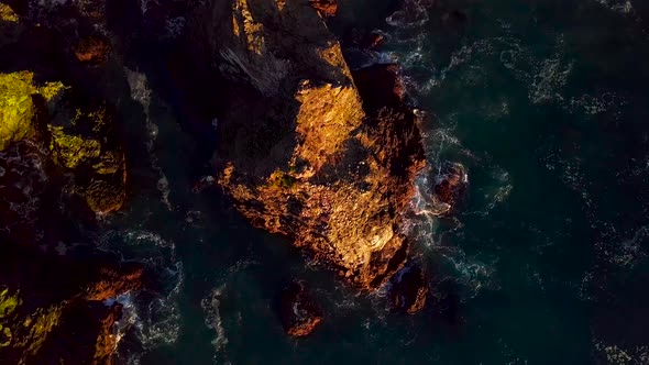 Rising wide aerial view of rocky cliffs and foaming ocean waves at sunset in Big Sur California