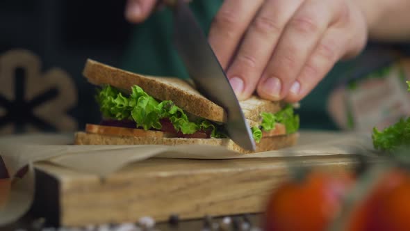 Man Cuts Club Sandwich in a Half By Sharp Knife on a Wooden Board at the Kitchen