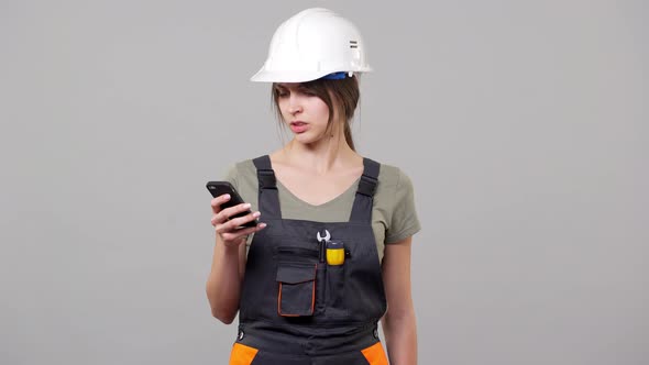 Portrait of Forewoman Builder in Helmet and Jumpsuit Smiling and Using Smartphone for Scrolling