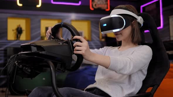 A Teenage Girl Wearing Virtual Reality Glasses Sits in a Car Driving Simulator