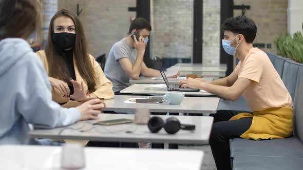 Side View of Concentrated Young Man in Face Mask Using Laptop with Female Friends Chatting at Front